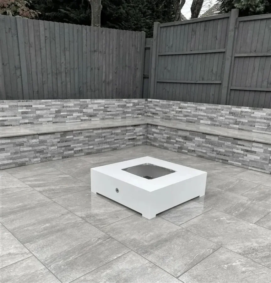 A modern outdoor patio with gray stone tile flooring and gray brick walls. A sleek, square white fire pit is centered on the patio. The background shows a tall wooden fence with trees behind it.