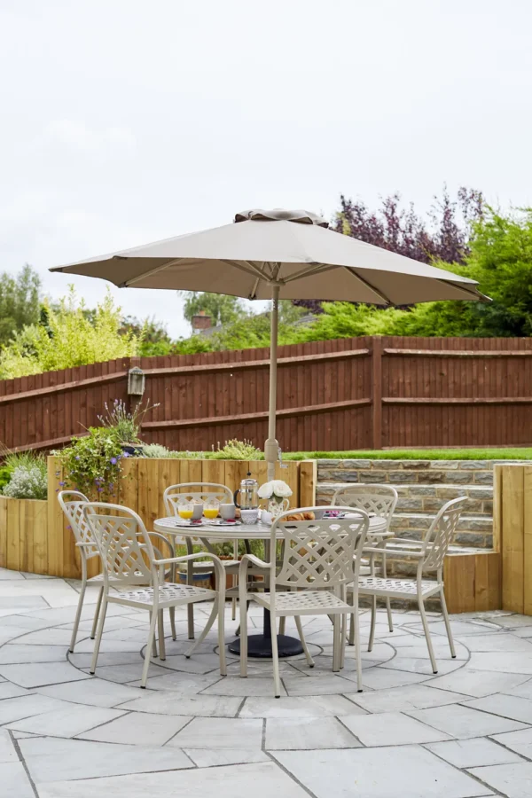 A patio with a round table and five chairs under a beige umbrella. The table is set with drinks and snacks. The area is surrounded by wooden fencing, plants, and a stone wall, with trees in the background.