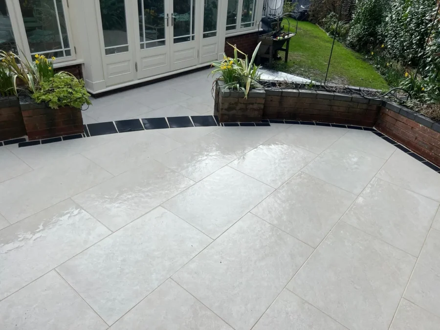 The expansive outdoor patio showcases Quartz White Outdoor Porcelain Paving Tiles, sized at 600mm x 600mm x 20mm. Two brick planters filled with greenery border the edge, and a glass door in the background opens into a building. Beyond the patio, a grassy area with more plants can be seen.