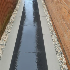 A narrow path of glossy black tiles bordered by Kandla Grey Outdoor Porcelain Paving Planks (900x200x20mm) and light-colored gravel runs between a wooden fence and a brick wall.