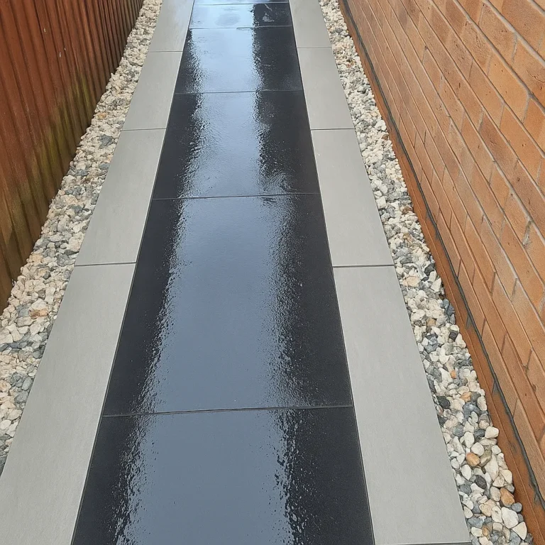 A narrow path of glossy black tiles bordered by Kandla Grey Outdoor Porcelain Paving Planks (900x200x20mm) and light-colored gravel runs between a wooden fence and a brick wall.