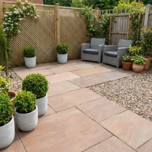 A small patio garden with Autumn Brown Indian Sandstone Paving Slabs (600x900mm), gravel, potted plants, green shrubs, and two gray wicker chairs beside a wooden lattice fence and gate.
