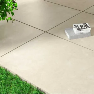 Close-up of an Aosta White Porcelain patio, sized 600mm x 900mm per tile, featuring natural paving next to lush green grass. A small, potted plant is visible in the top left corner. Two stacked books rest on the tiles in the top right.