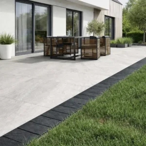 Modern patio features large light gray tiles with a border of Black Anthracite Outdoor Porcelain Cobble Setts (100x200x20mm), green grass, and a dining table for four near glass doors. Potted plants add greenery to this contemporary outdoor space.