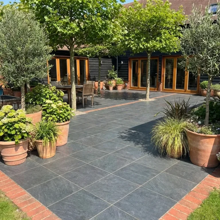 A sunlit patio features County Black Anthracite Outdoor Porcelain Paving Tiles (600mm x 600mm x 20mm) with a red brick border, surrounded by potted trees. Wooden chairs and tables sit near the building’s large glass doors and windows.