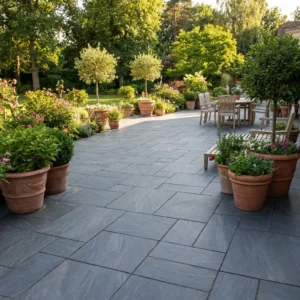 A spacious patio paved with Anthracite Black Outdoor Porcelain Paving Tiles (Mixed Sizes), lined with potted plants and small trees. A wooden dining set sits on one side, surrounded by lush greenery and a garden backdrop.