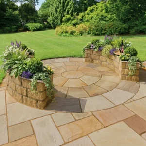 A Camel Buff 2.4 Meter Circle - 22mm Calibrated stone patio with a low, curved wall and built-in planters overflows with greenery and flowers, surrounded by a manicured lawn and dense trees.