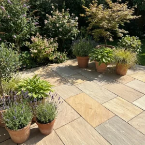 Potted lavender, hostas, and grasses sit on a patio paved with Camel Buff Indian Sandstone Slabs (600mm x 900mm), surrounded by lush shrubs and trees in bright garden sunlight.