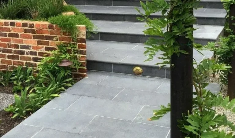 A set of elegant Kandla Grey Sandstone Bullnose Steps, reminiscent of cobble setts, winds upward through lush greenery. On the right, a post adorned with climbing plants; on the left, a brick wall with vibrant plants and flowers at its base. A small decorative shell graces one step.
