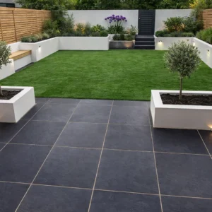 Modern backyard features large Black Limestone Sawn Edge 600x600mm tiles, rectangular raised white planters, small trees, manicured lawn, wooden fence, garden lighting, and steps leading to a higher area with more plants and flowers.