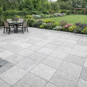 A patio with Silver Grey Granite 600mm x 600mm x 20mm tiles features a table and four chairs, surrounded by green grass, vibrant flower beds, and dense foliage.