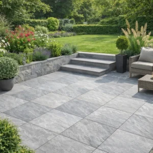 A modern patio features Kandla Grey Outdoor Porcelain Paving Tiles (600mm x 600mm x 20mm), bordered by lush plants and vibrant flowers. Wicker chairs with cushions and a coffee table rest near stone steps that lead to a grassy lawn.
