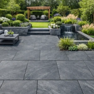 Modern backyard patio featuring County Black Anthracite Outdoor Porcelain Paving Tiles (600x600mm), a cozy seating area, coffee table, lush plants, a small waterfall feature, and a pergola with extra seating in the background.
