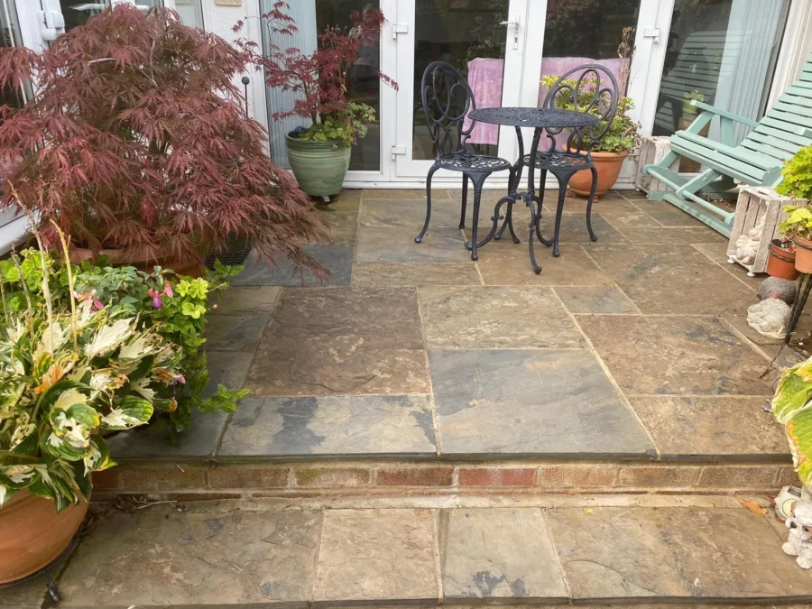 A patio with stone tiles features a black wrought iron table and two chairs. Surrounding the area are various potted plants, including a red-leafed tree and lush greenery. A pink towel hangs on a chair, and a wooden bench is in the background.