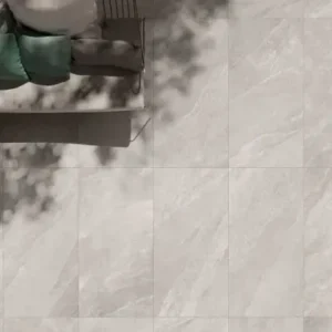 Top view of County Ivory Outdoor Porcelain Paving Tiles (600mm x 900mm) with sunlight casting leafy shadows; a sofa with green and white cushions is partly visible in the upper left corner.
