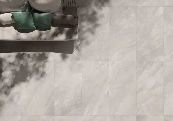 Top view of County Ivory Outdoor Porcelain Paving Tiles (600mm x 900mm) with sunlight casting leafy shadows; a sofa with green and white cushions is partly visible in the upper left corner.