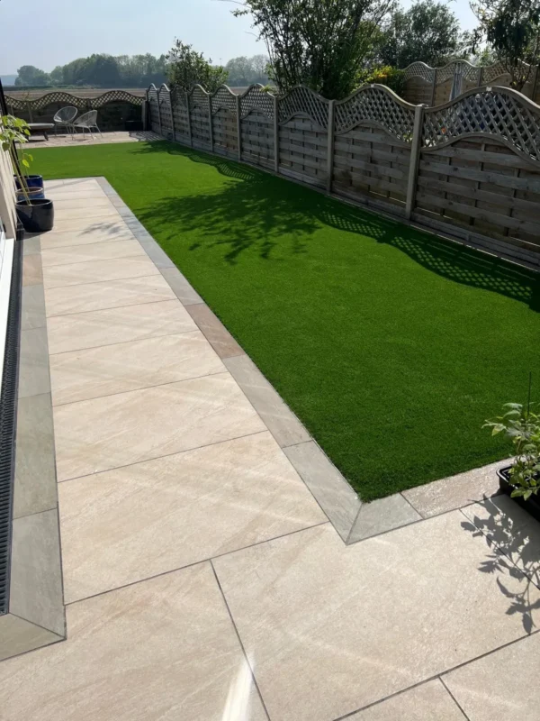 A sunny backyard featuring neatly landscaped surroundings showcases a paved patio made from County Crema Outdoor Porcelain Paving Tiles (600mm x 900mm x 20mm) alongside a rectangular patch of artificial grass. The garden is enclosed by wooden fencing, with potted plants and outdoor seating visible in the distance.