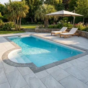 A small rectangular backyard pool with curved steps, surrounded by county light grey porcelain paving tiles. Two lounge chairs with cushions sit under a large umbrella, with palm trees and lush greenery in the background.