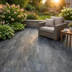 A stone-tiled patio featuring Earthcore Black Outdoor Porcelain Paving Tiles (600x900x20mm), with a wicker armchair and wooden side table, surrounded by lush greenery and flowers, basking in sunset light in a landscaped backyard.