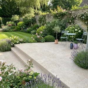 A stone patio featuring Egyptian limestone paving 600 x 900 and steps leads to a lush garden with blooming flowers, shrubs, and a stone wall. On the patio, there’s a small round table with two metal chairs and decorative vases.