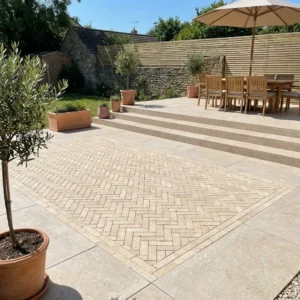 A patio paved with Sinai Pearl Washed and Tumbled Limestone Setts (200x50x40mm) in a herringbone pattern, potted olive trees, a wooden dining table under an umbrella, steps, and a wooden fence with lush greenery behind.