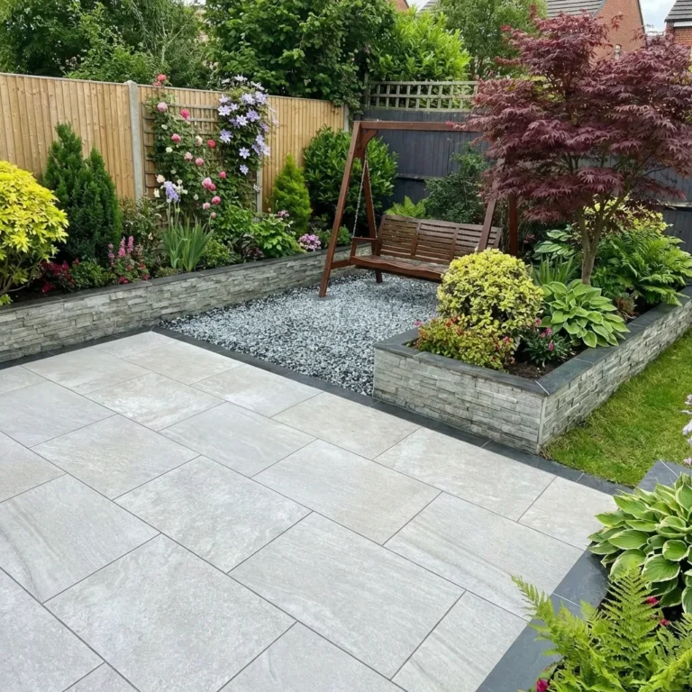 A neatly landscaped garden featuring a swing bench on a gravel area, surrounded by raised flower beds with various shrubs and flowers, adjacent to an elegant Everest pearl porcelain paving 600 x 900 light grey tiled patio.