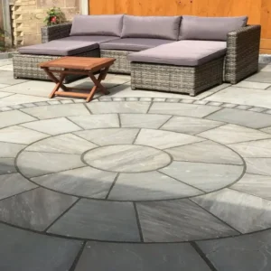 A patio featuring a Kandla Grey 3 Meter Circle stone pattern on the ground showcases a wicker outdoor sectional sofa with gray cushions and a small wooden folding table. The backdrop includes a wooden fence and part of a house wall.