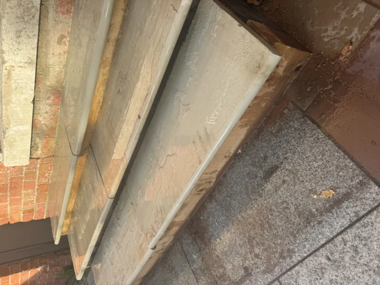 Fossil Mint Sandstone Bullnose Steps 900x350x35mm appear wet, showing water tracks and some dirt. Bordered by a brick wall, the steps lead down to stone paving, with sunlight highlighting their textured surfaces.