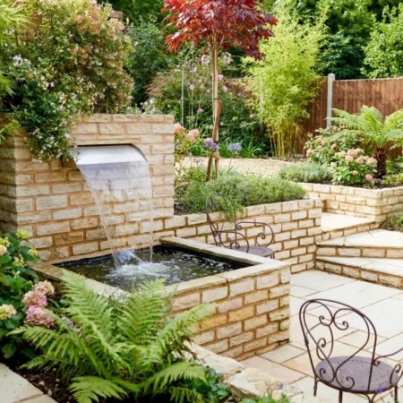 A small landscaped garden with a fossil mint sandstone wall, brick water feature, cascading stream, lush green plants, flowers, two metal chairs, and stone steps leading to a higher level.