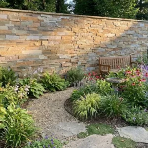 A curved gravel path leads to a wooden bench set before a Garda Buff Stone Cladding wall, surrounded by lush green plants and colorful flowers for a tranquil garden atmosphere.