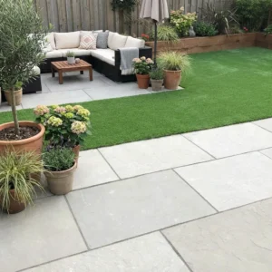 A modern backyard patio features Grey Limestone Sawn Edges (22mm Calibrated, 600x900mm) tiles, potted plants, a grassy area, black outdoor sofa with cushions, a coffee table, umbrella in the corner, wooden fence and raised garden beds.