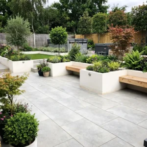 Modern backyard garden with large HS Grey Porcelain paving tiles, white raised planters filled with green shrubs and small trees, wooden benches, potted plants, and a grassy area; surrounded by a wooden fence and lush foliage.