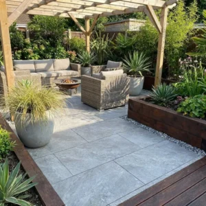 A modern patio features Himalayan Grey Outdoor Porcelain Paving Tiles (600x900x20mm), rattan furniture, a fire pit, wooden pergola, and lush potted plants in raised beds, all surrounded by a garden.