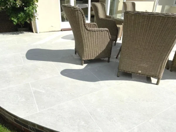 A sunlit patio with light gray tiles, featuring three wicker chairs and a round table. The chairs cast distinct shadows on the tiles, and greenery is visible in the background near a glass door.