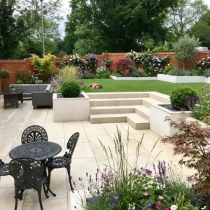 A landscaped garden with a paved patio featuring HS Beige porcelain paving 600 x 900, black metal table and chairs, raised flower beds, steps to a grassy lawn, and a dog on the grass. Shrubs, flowers, and trees surround the area.