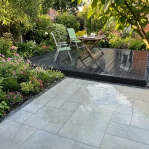 A garden patio features HS Grey Outdoor Porcelain Paving Tiles in mixed sizes, leading to a raised wooden deck with two chairs and a table, all surrounded by lush green plants and colorful flowers.