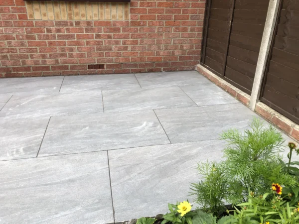 A cozy patio features large Grey Anthracite Porcelain tiles measuring 600mm x 900mm, bordered by a red brick wall and a brown wooden fence. In the foreground, small green plants with yellow flowers enhance the tranquility of this serene space.