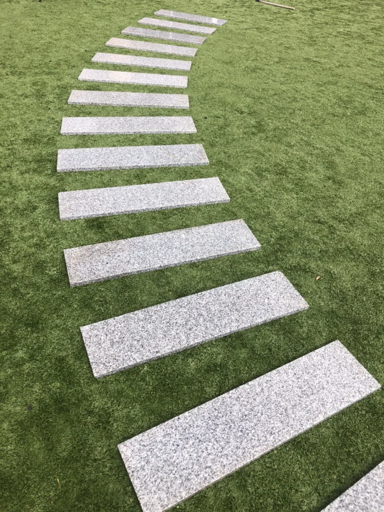 A curved row of Silver Grey Granite G603 Planks (200mm x 900mm) is arranged on green artificial grass, creating a visually striking pattern.