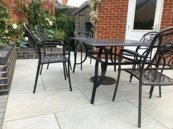 A charming patio scene featuring Hs Grey Outdoor Porcelain Paving Sample. In the background, a white door, a brick wall, and a lush garden flourish with blooming flowers and greenery.