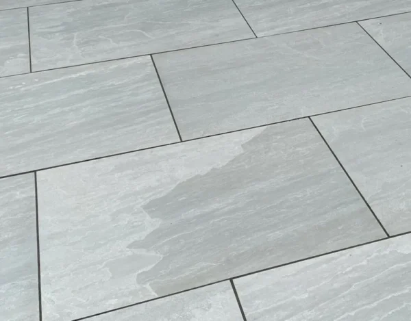 Close-up of a dry outdoor patio featuring Kandla Grey Outdoor Porcelain Paving sample. A small section of one tile looks darker, as if wet. The enduring elegance of these tiles captivates the eye and elevates the tranquil ambiance.