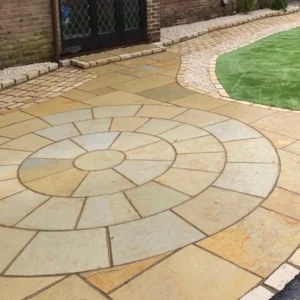 A Yellow Limestone 3 Meter Paving Circle forms a tan and beige stone patio, bordered by a curved brick path and artificial grass near glass double doors.