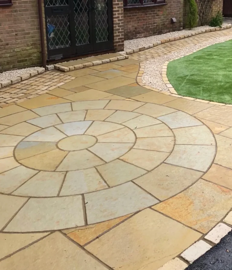 A Yellow Limestone 3 Meter Paving Circle forms a tan and beige stone patio, bordered by a curved brick path and artificial grass near glass double doors.