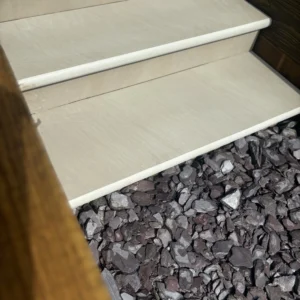 A close-up view of a staircase showcases Raj Green Honed Sandstone Bullnose Steps (900x350x35mm) that gracefully lead down to a bed of dark, rough stones. Wooden sides frame the staircase, while sunlight accentuates the textures of both the steps and stones.