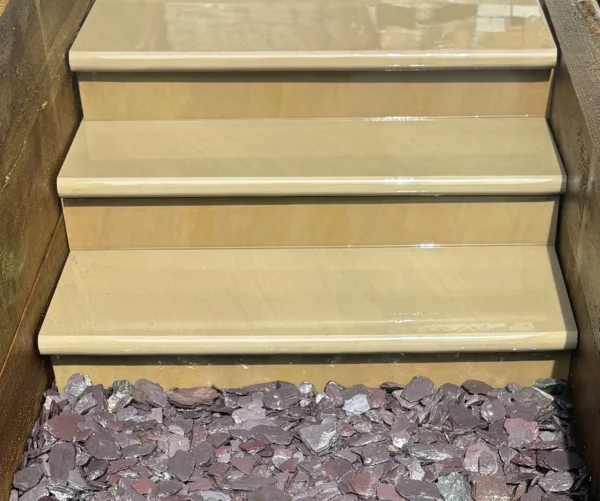 Four steps crafted from Raj Green Honed Sandstone Bullnose, each measuring 900x350x35mm and in a beige hue, lead down to a bed of purple slate rocks. The polished stone steps are bordered by wooden sides, and the sunlight casts a gentle glow across their surface.