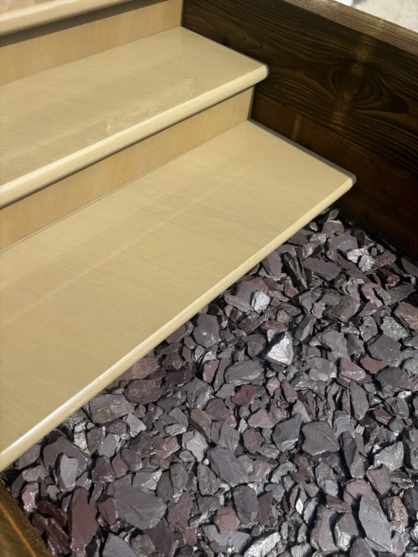 A set of Raj Green Honed Sandstone Bullnose Steps (900x350x35mm) with a wooden border leads down to a bed of purple and gray rocks, partially illuminated by sunlight.