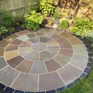 A circular stone patio crafted from raj green Indian sandstone features a stunning 3m mosaic pattern, nestled amidst lush plants and vibrant flowers. A wooden fence and rich greenery frame this serene outdoor haven.