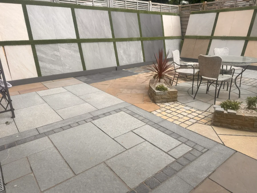 A modern patio with various stone textures and colors is showcased at the Natural Paving Store Showroom. It features an outdoor dining set and planters with decorative stones and plants, with walls lined by large, contrasting stone panels separated by green strips.