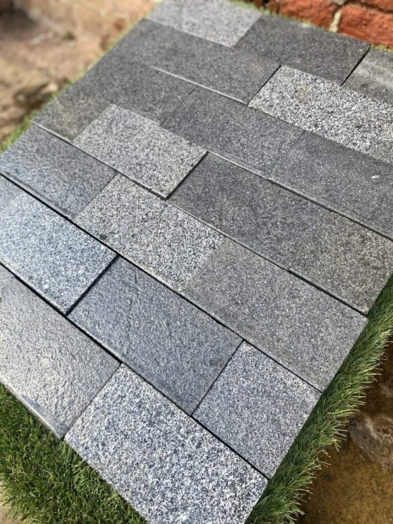 A close-up of a showcase featuring rectangular, textured gray tiles in a brick pattern on an artificial grass patch, seamlessly integrated with Blue/Black Granite Sawn Edge Setts (200mm x 100mm x 50mm) and set against a brick wall backdrop.