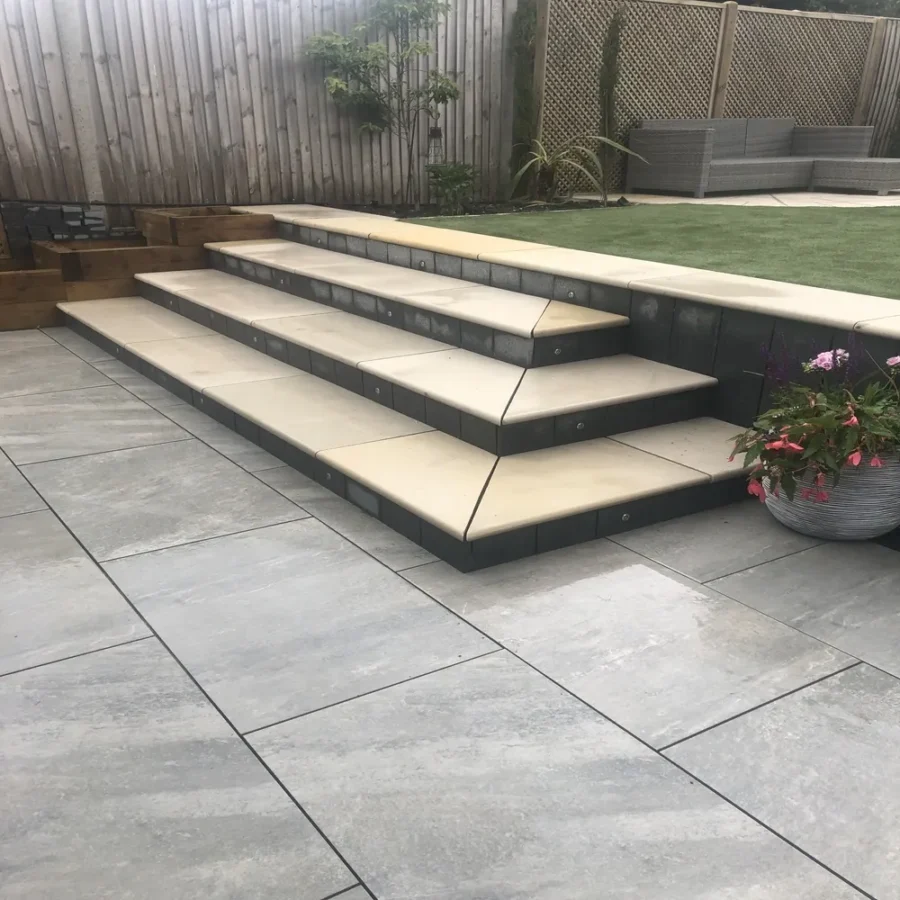 A contemporary garden featuring ivory mint sandstone bullnose steps that ascend to an artificial grass area. The patio showcases large tiles with a gray hue. Positioned to the right is a potted plant adorned with pink flowers, all enclosed within a wooden fence. The steps are crafted using the Ivory Mint Bullnose 900x350x35mm sample.