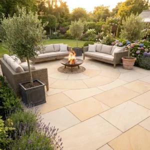 A stone patio paved with Ivory Mint Sandstone Honed 22mm Calibrated 600x900mm, featuring cushioned outdoor sofas arranged around a fire pit and surrounded by potted trees and blooming flowers at sunset.